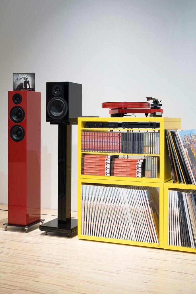 Red turntable on top of a yellow shelf filled with books next to a floorstanding speaker and a bookshel speaker on a stand